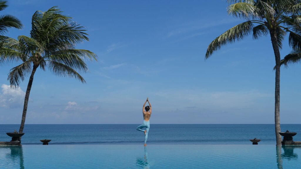 A guest doing yoga at the edge of the infinity pool at The Legian Seminyak, palms and the sea behind