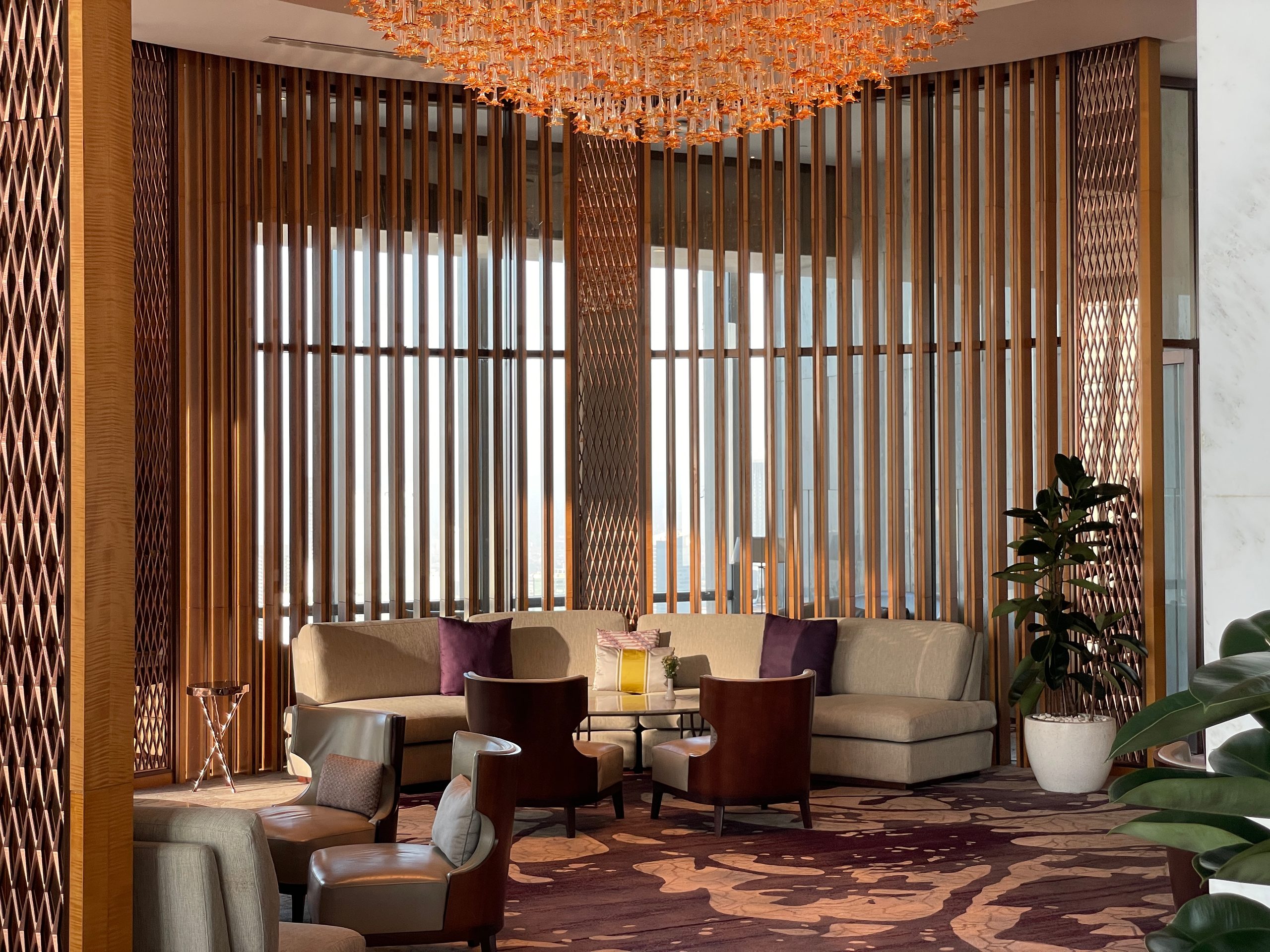The seating area beneath the orange glass chandelier, screened in timber, with the city through the slats — The Westin Jakarta