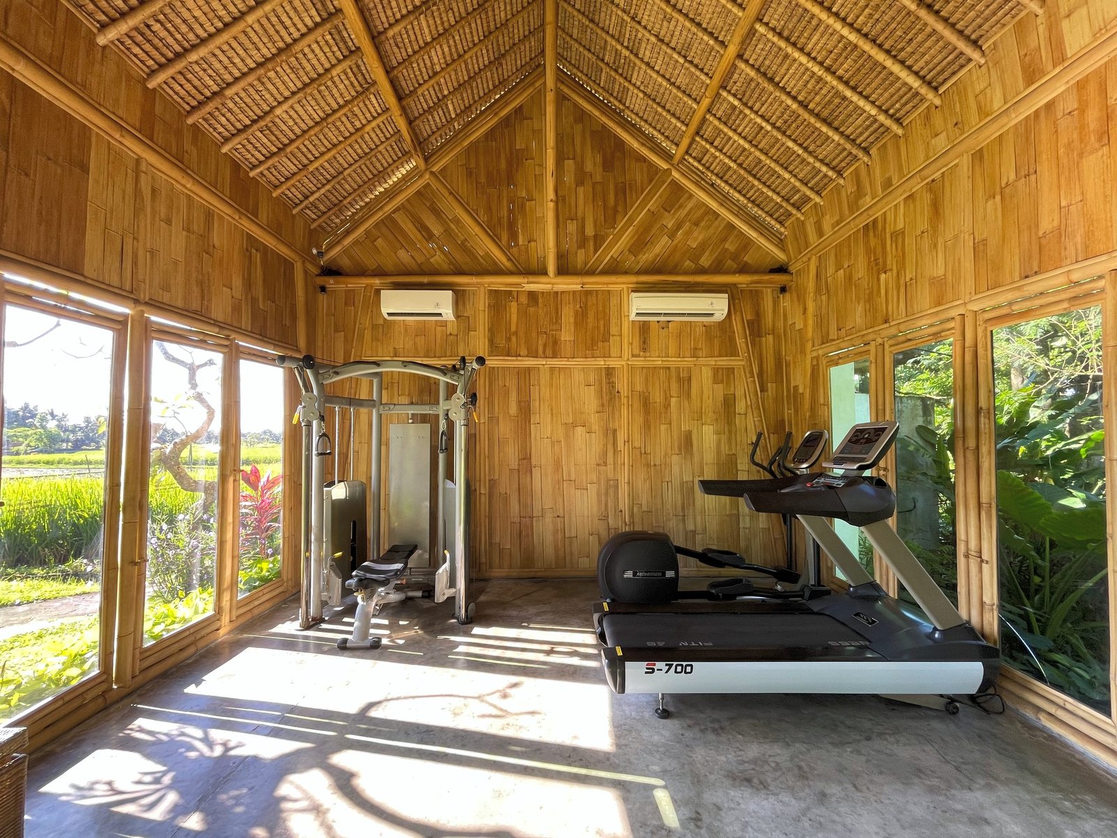A treadmill and cable machine inside the bamboo gym at Visesa Ubud, rice fields through the windows