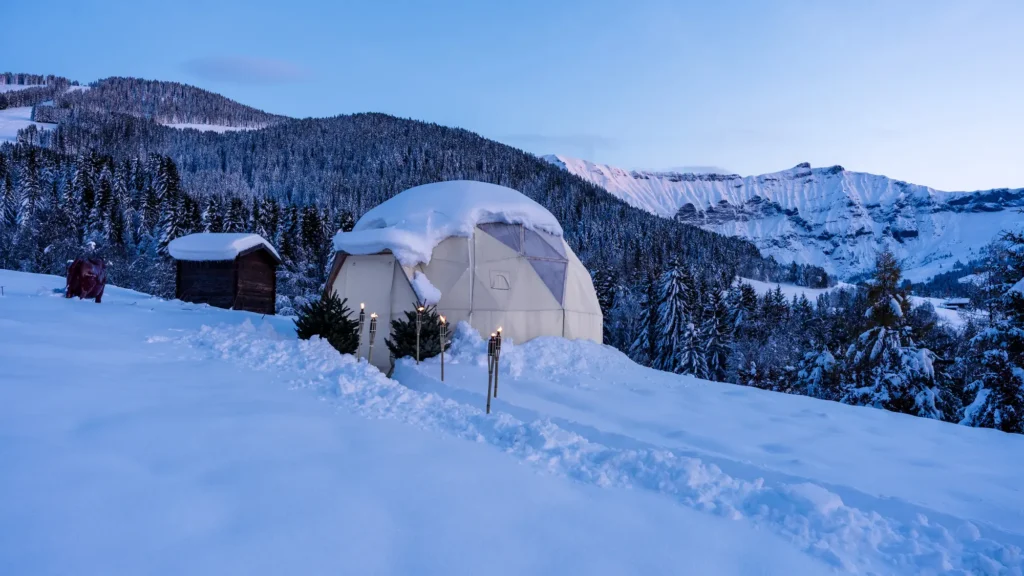 A white geodesic dome in deep snow on a mountainside above a forested valley
