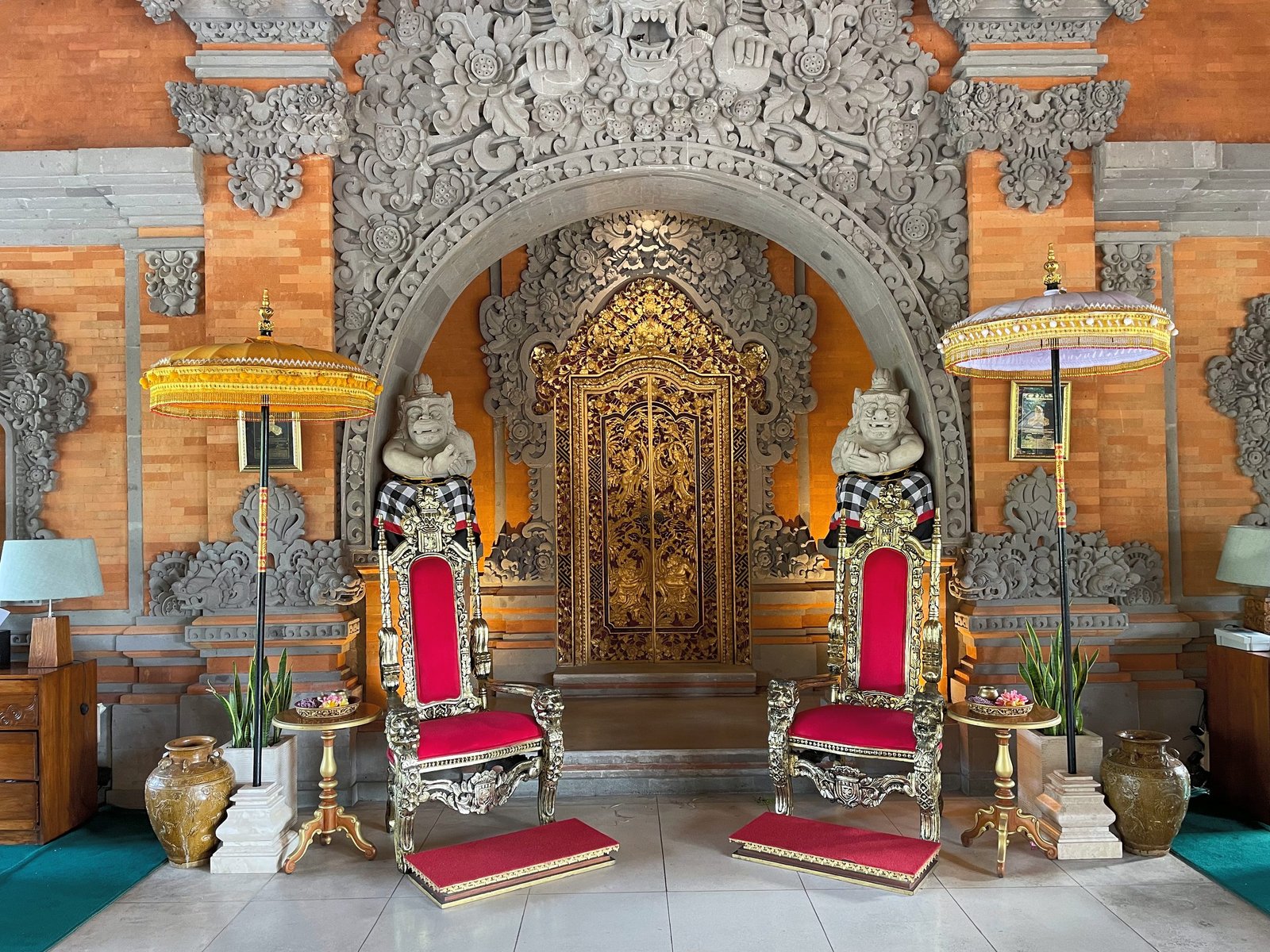 A gilded carved doorway set in a brick and stone wall, flanked by red thrones and parasols, in the lobby of Visesa Ubud