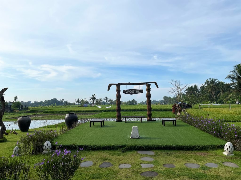A carved wooden gateway and stone benches on a lawn, with working rice fields behind, at Visesa Ubud