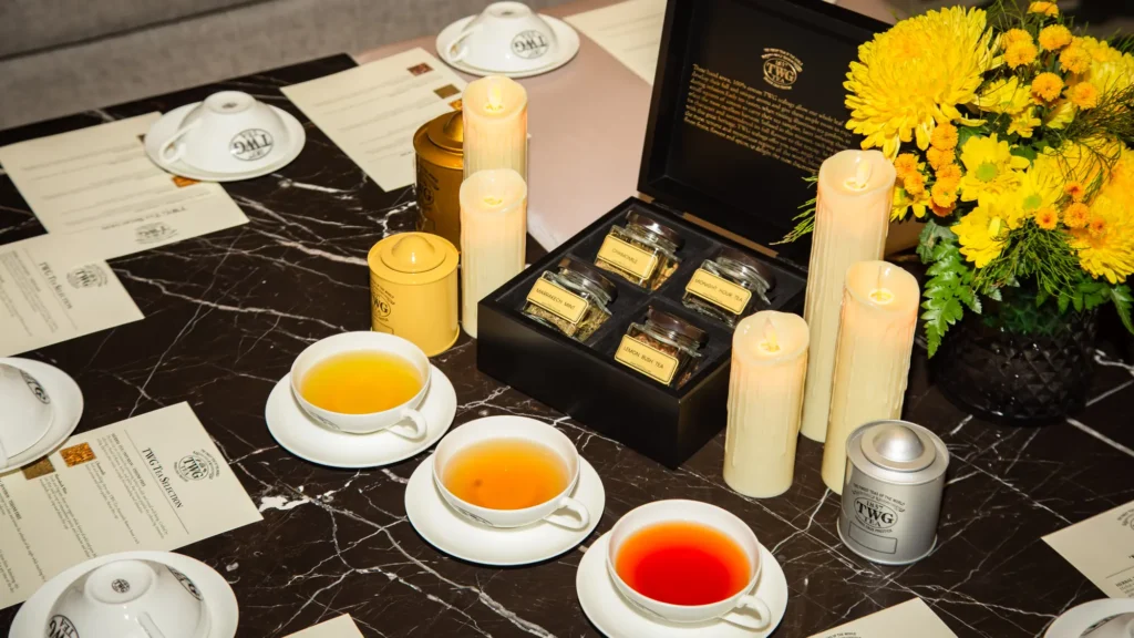 Three cups of TWG tea beside a boxed tea selection, candles and yellow chrysanthemums on a black marble table