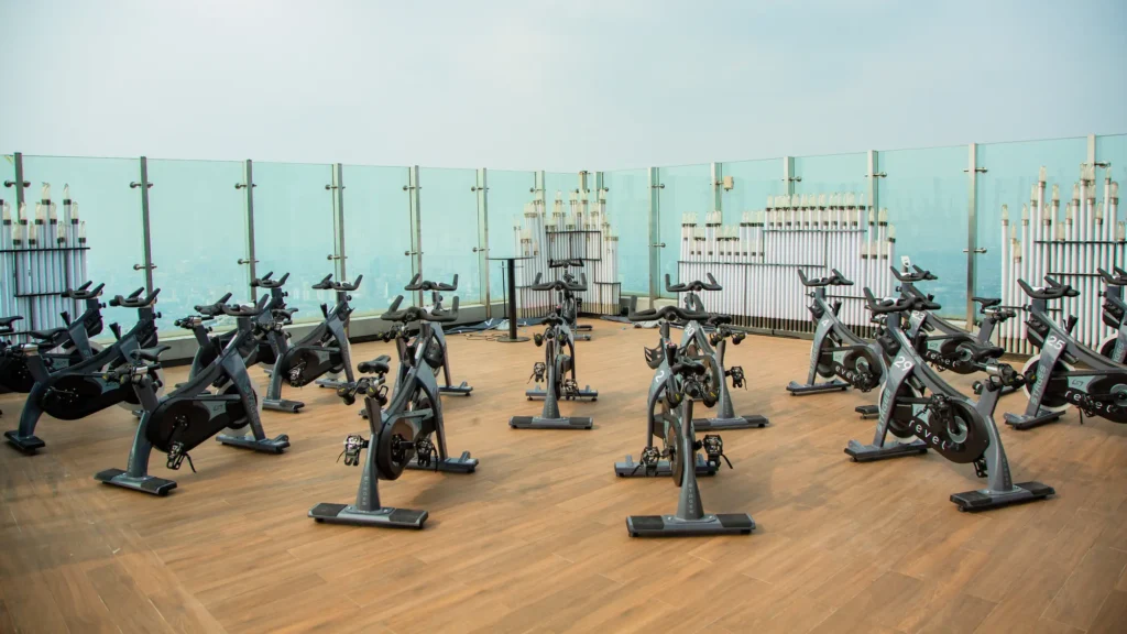 Rows of indoor-cycling bikes on a wooden rooftop deck behind a glass balustrade, with the Jakarta skyline in haze