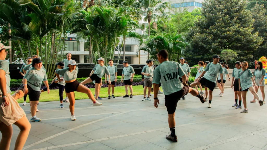 Runners in matching sage-green T-shirts doing a warm-up routine on a paved forecourt lined with palm trees