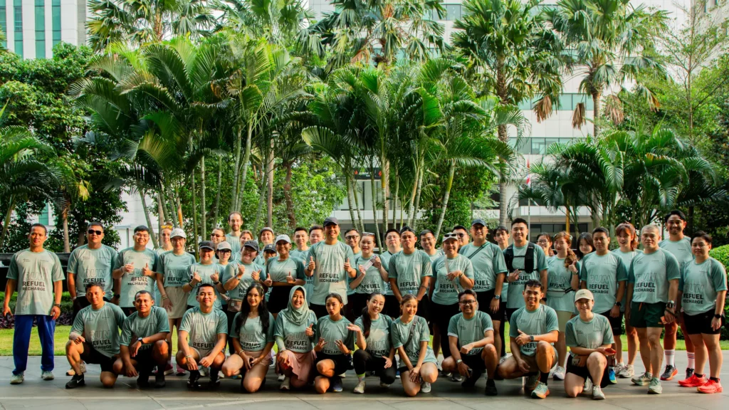 A large group of runners in matching sage-green Run, Refuel, Recharge T-shirts posing in front of palm trees in Jakarta