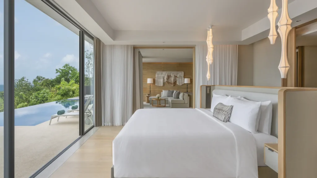 Bedroom with a white bed, sculptural pendant lights and sliding glass doors onto an infinity pool