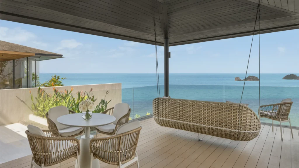 Covered wooden deck with a hanging rattan swing seat, dining table and chairs overlooking the sea