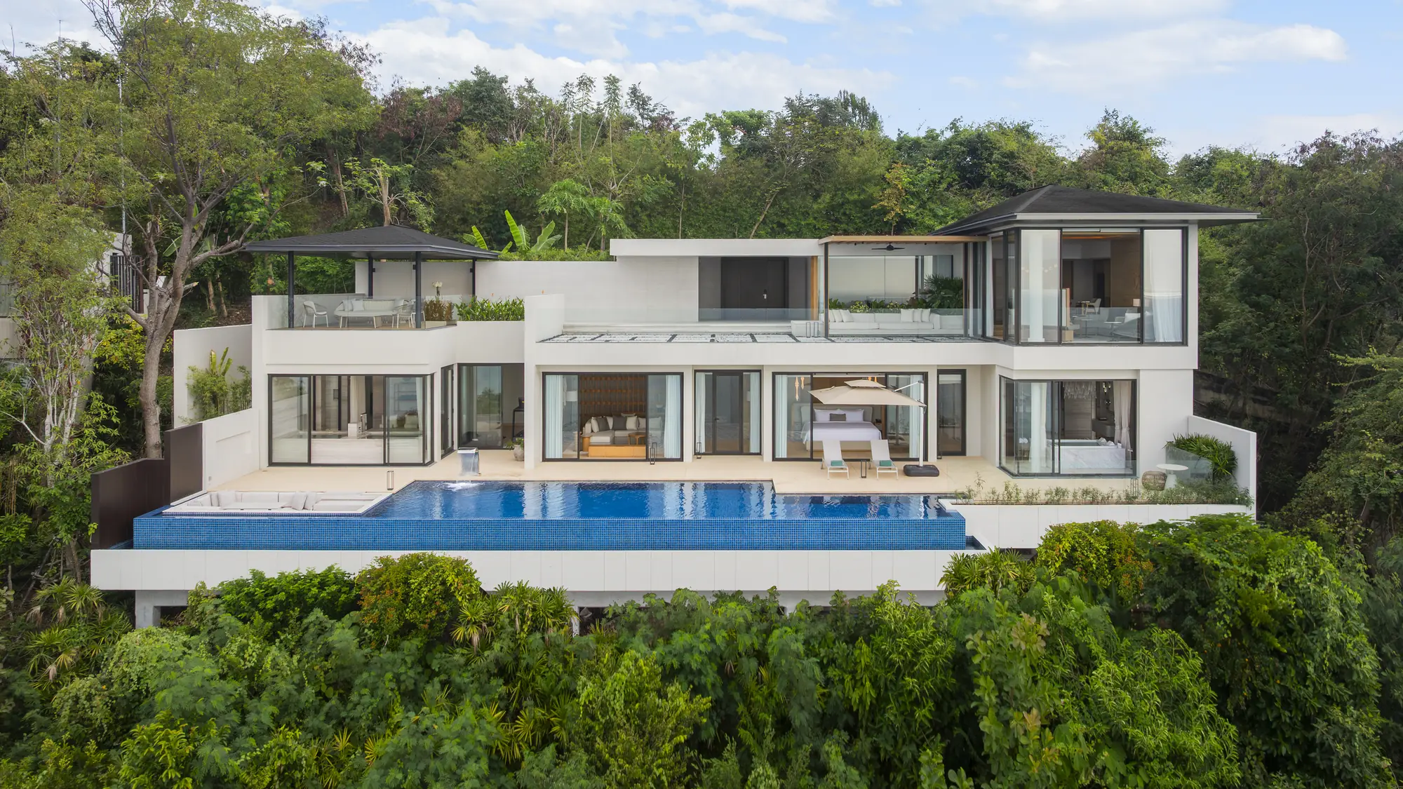 White two-storey hillside villa with a long blue infinity pool, surrounded by tropical forest