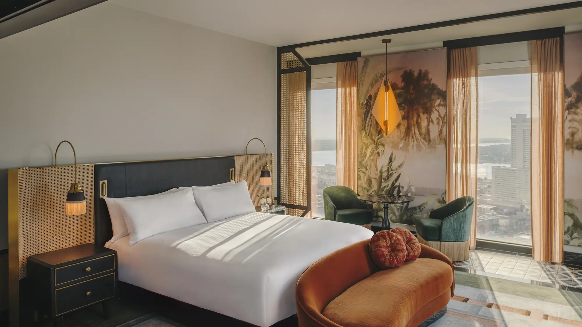 Hotel bedroom with a cane headboard, rust velvet sofa and floor-to-ceiling windows over the New Orleans skyline