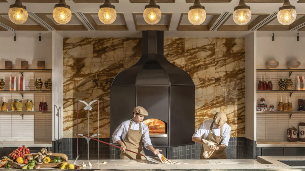 Two chefs in flat caps working at a black wood-fired oven set into a marble wall