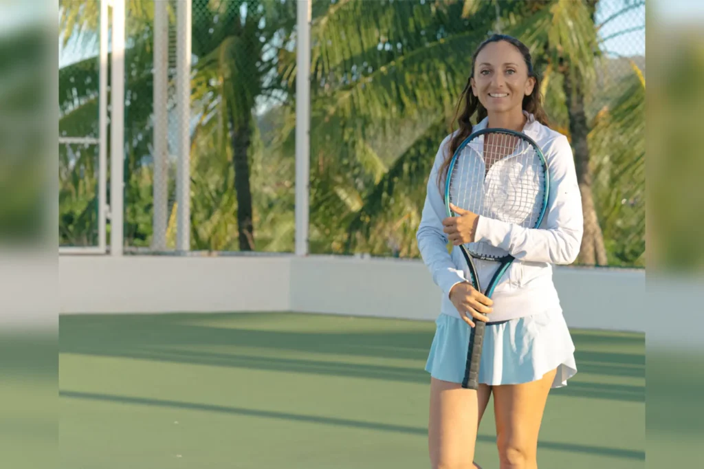 A smiling woman in tennis whites holding a racket on a court framed by palms