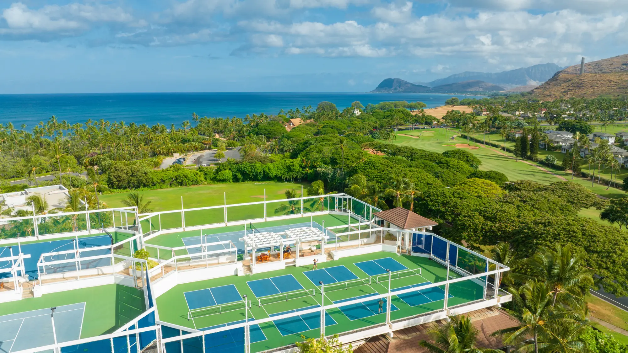 Aerial view of fenced tennis and pickleball courts among palms and golf greens, with the ocean beyond