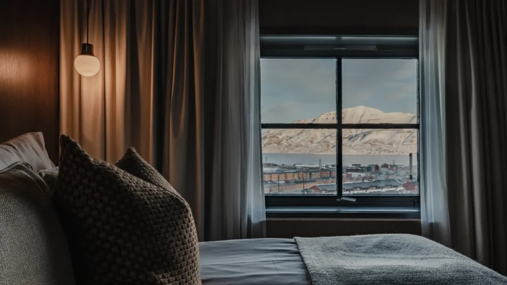 Dark guest room with a bed and a window framing snowy mountains over Longyearbyen
