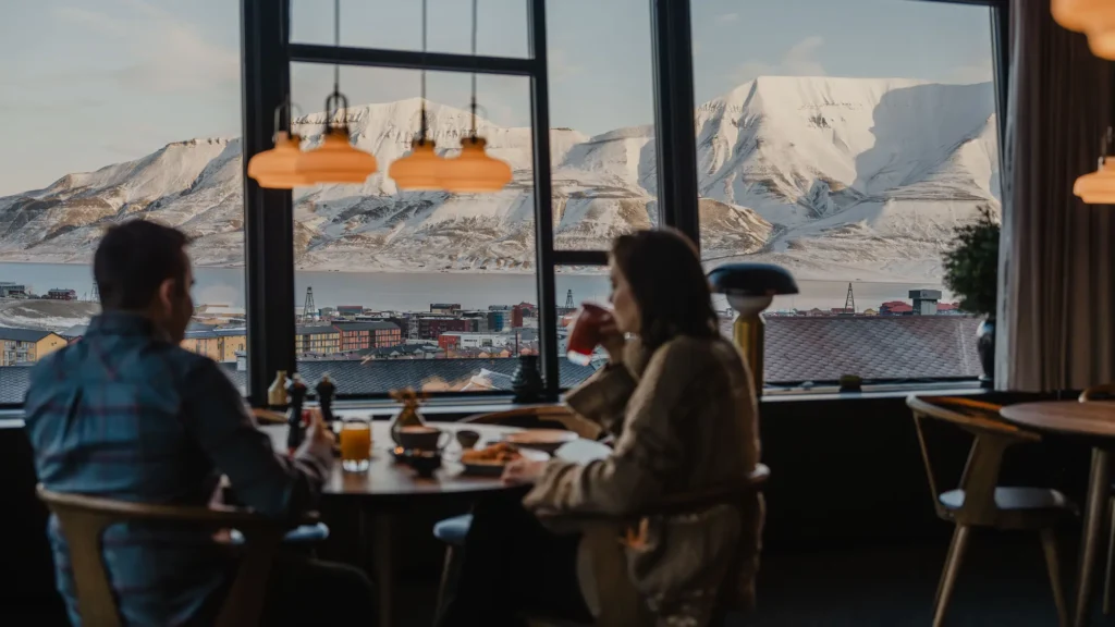 Two guests at breakfast by a picture window overlooking Longyearbyen and snow-covered mountains
