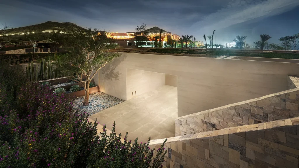 Stone-walled entrance court lit at night, with desert planting and hills beyond
