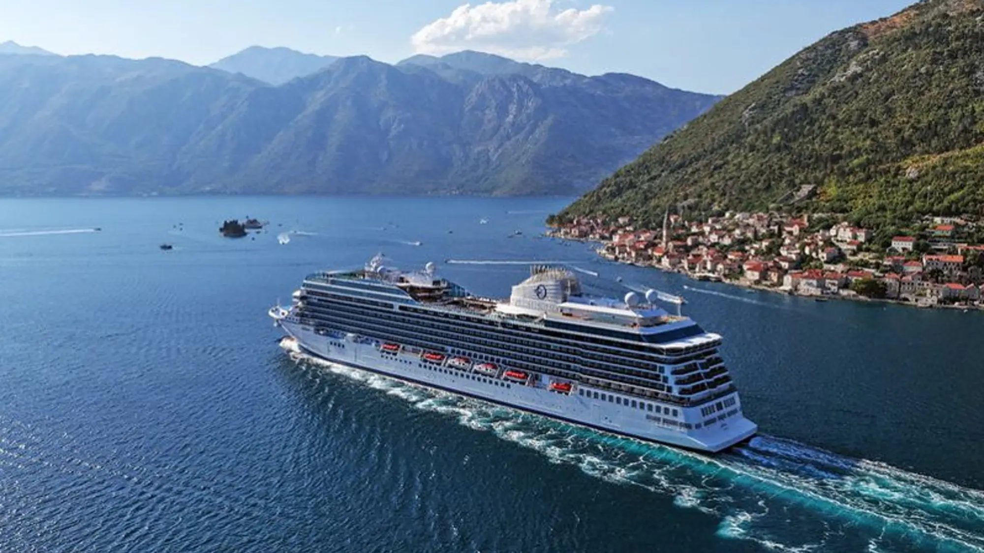 White cruise ship sailing past a red-roofed waterfront town beneath mountains in the Bay of Kotor