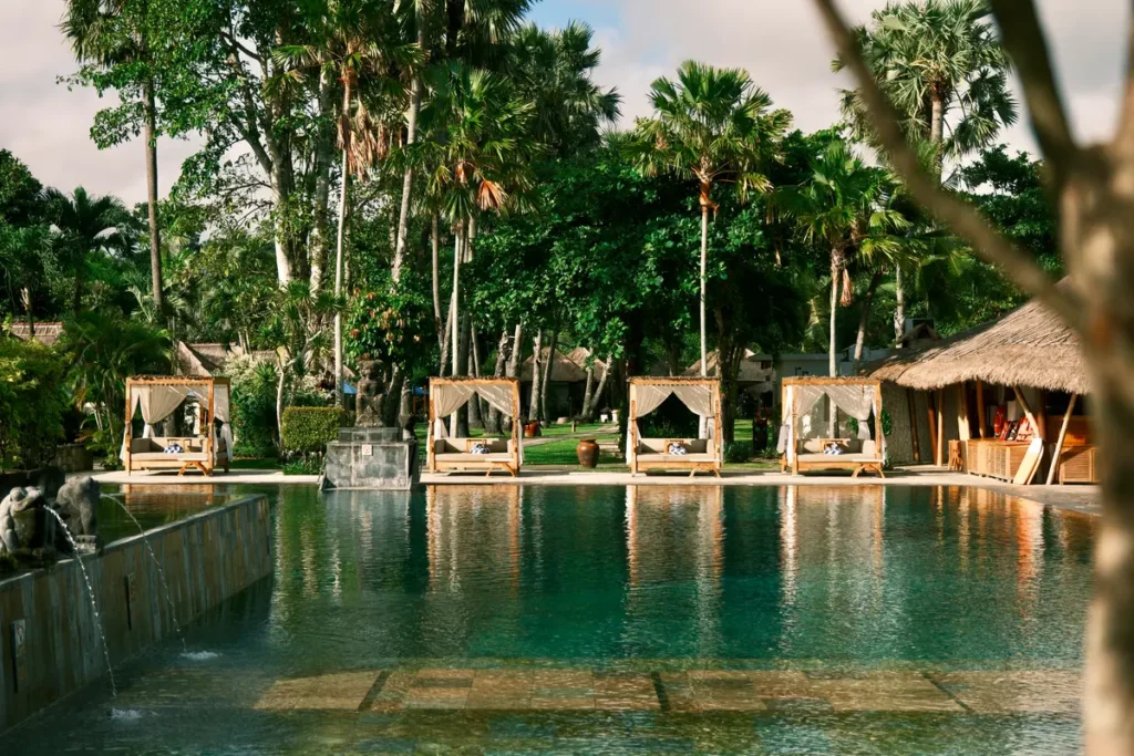 The pool at Jimbaran Puri, A Belmond Hotel, lined with daybeds and thatched pavilions under palms
