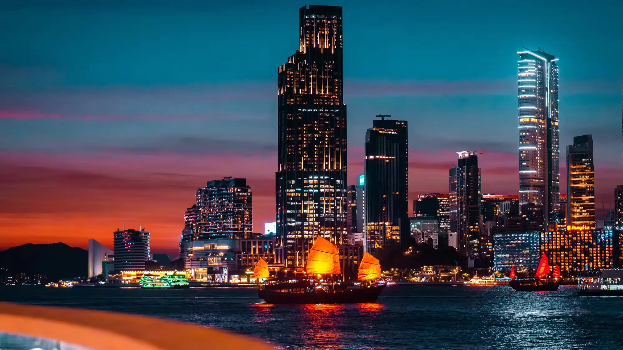 Hong Kong harbour at dusk with a junk under illuminated sails in front of the Rosewood Hong Kong tower