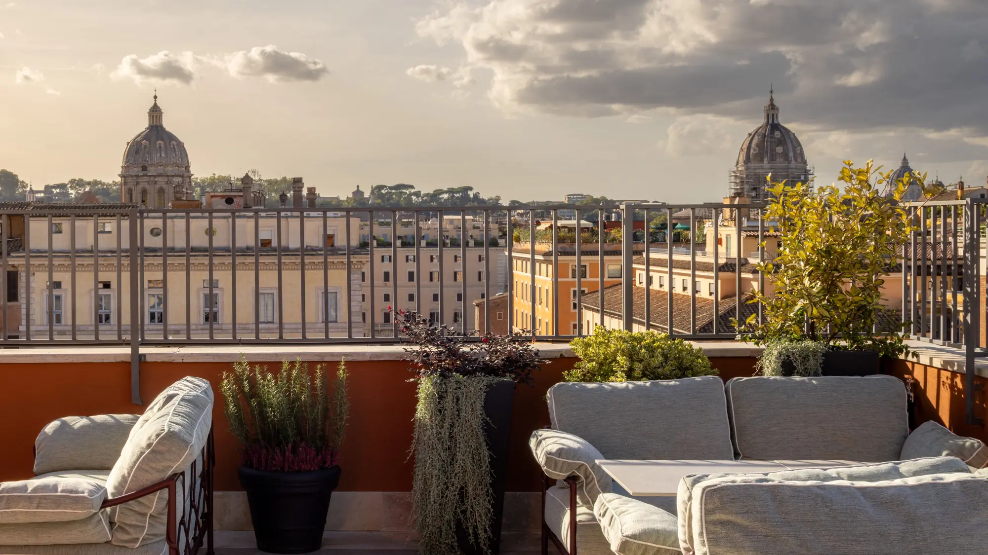 Rooftop terrace with sofas and potted plants overlooking Rome's church domes under a cloudy sky