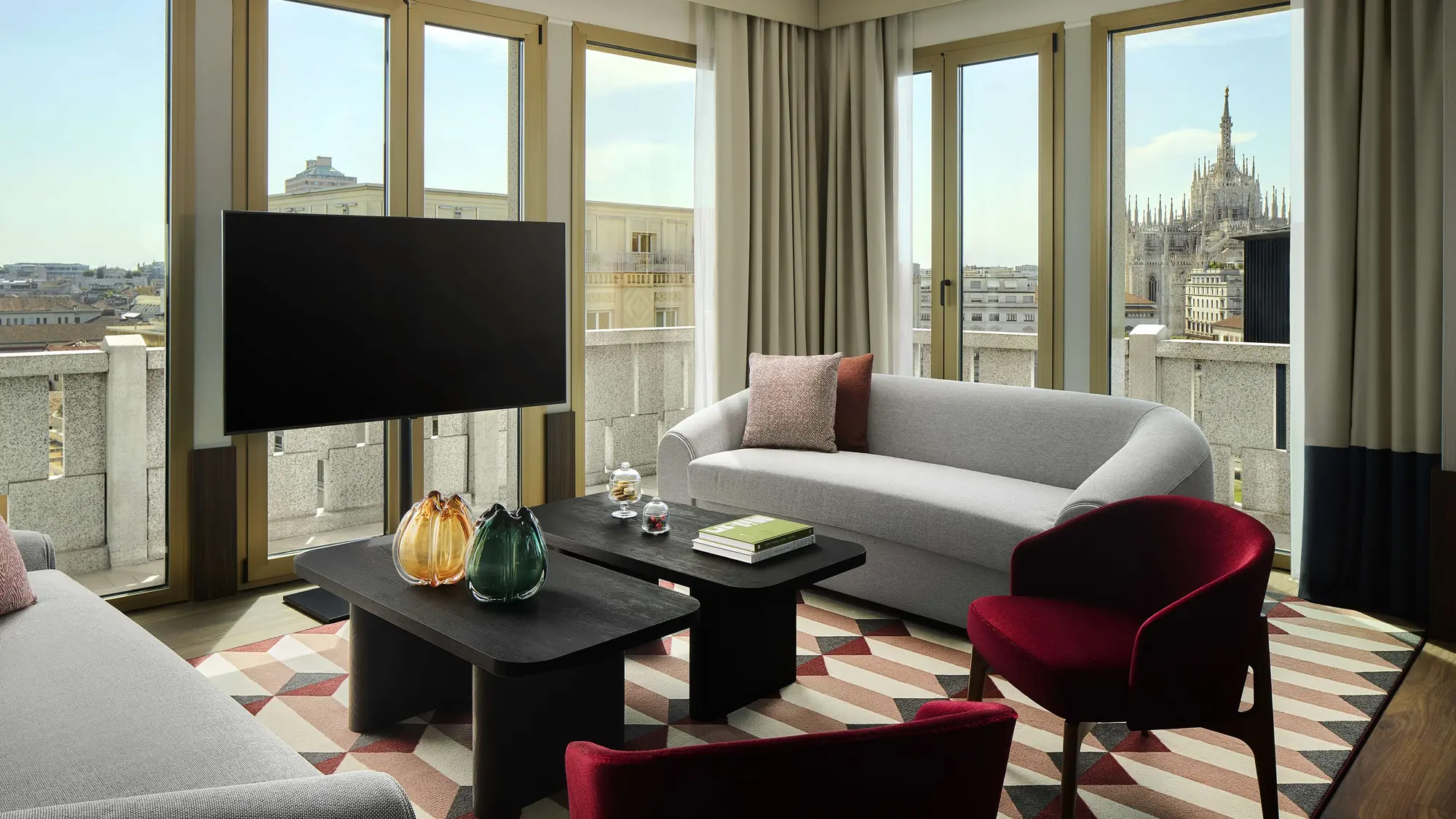 Living room of the Presidential Suite at Tivoli President Milano with red armchairs, a grey sofa and the spires of Milan Cathedral through the window
