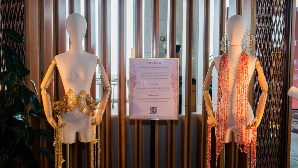 Two wooden mannequins wearing gold and coral-red jewellery either side of a TULOLA display card
