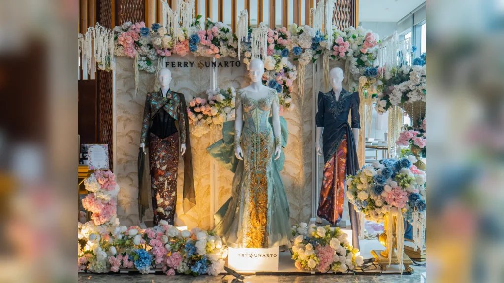 Three mannequins in embellished Indonesian couture gowns surrounded by pink, white and blue flowers under a Ferry Sunarto sign