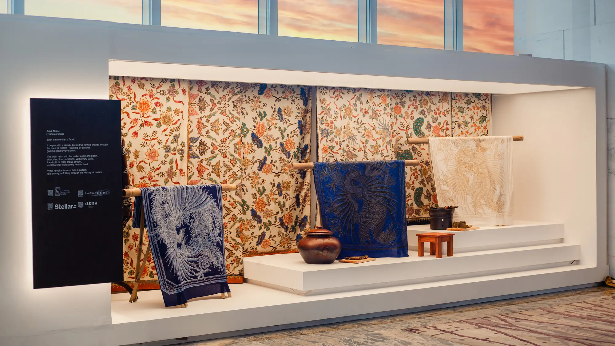 Lit display alcove of batik cloths on bamboo rails and clay pots beneath windows with a sunset sky at The Westin Jakarta